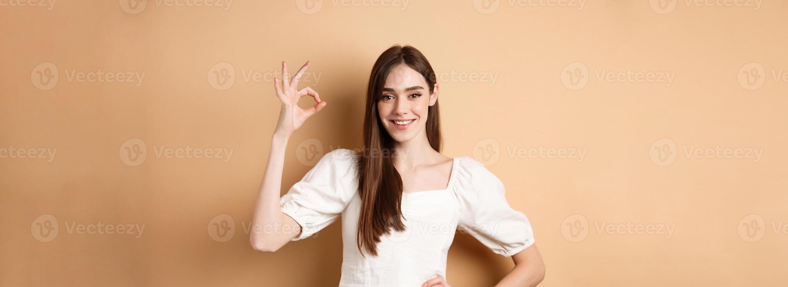 Very good. Smiling cute woman in white blouse showing okay sign in approval, like and praise excellent choice, standing satisfied on beige background photo