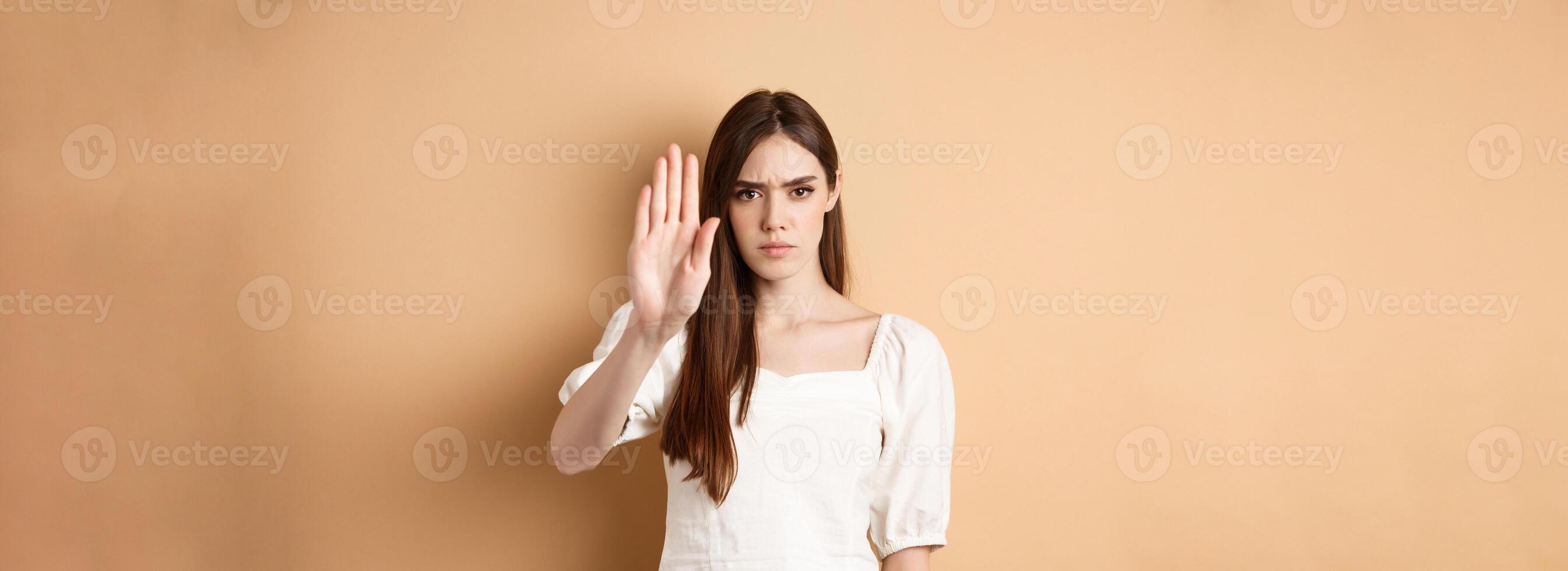 Nope stop right there. Serious and confident woman stretch out hand to forbid something, frowning and saying no, disagree and reject bad offer, standing on beige background photo