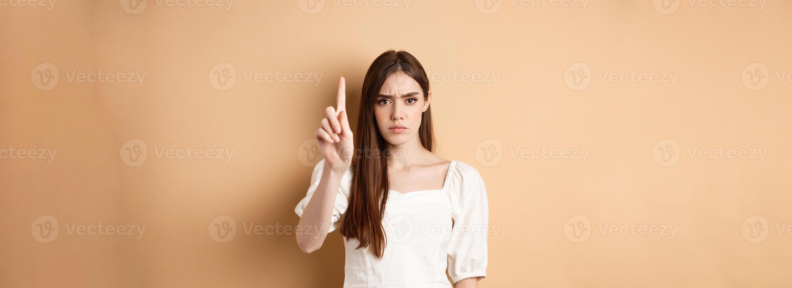 Rule number one. Serious frowning woman showing finger to scold person, look disappointed and angry, standing on beige background photo