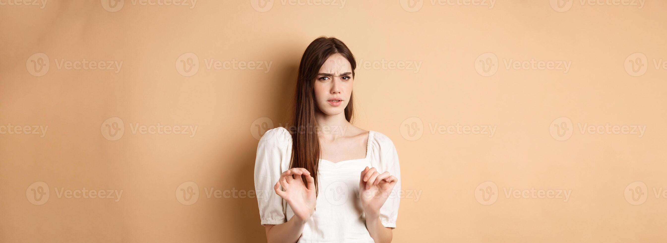 Stay away from me. Disgusted young woman frowning and looking with aversion at something stinky, rejecting disgusting thing, standing on beige background photo
