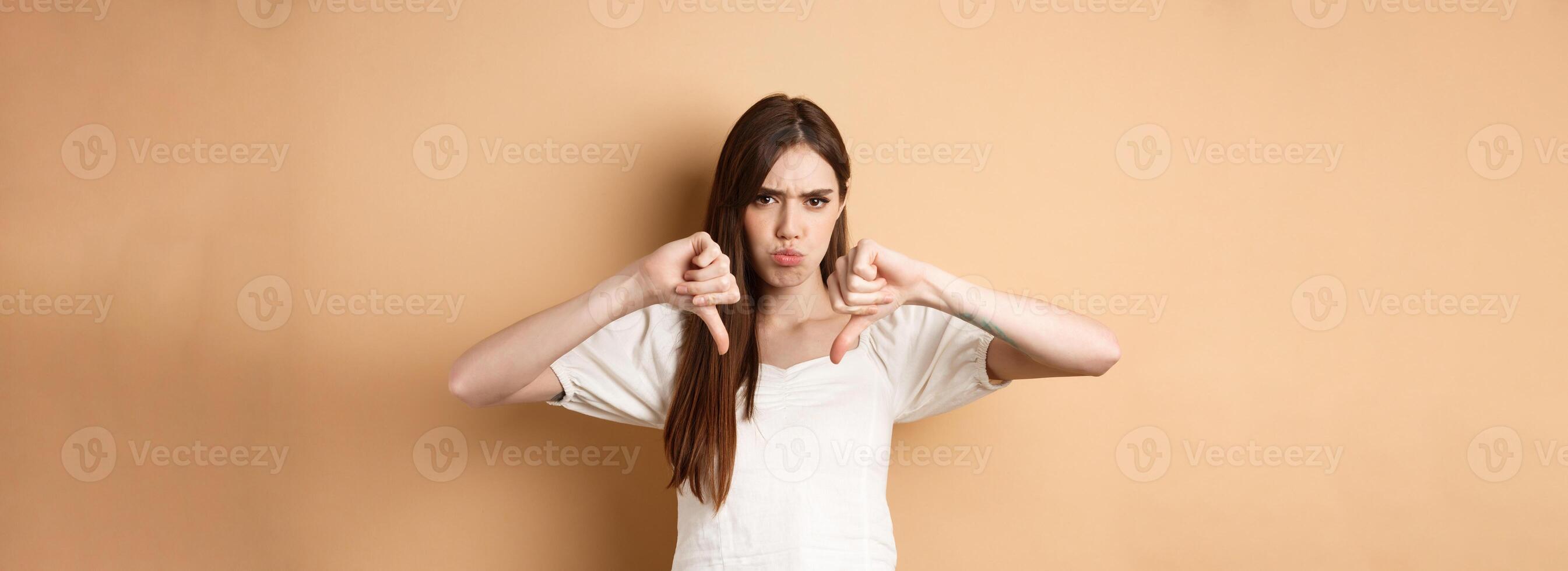 Absolutely no. Disappointed young woman frowning and showing thumbs down, express dislike and negative emotions, bad feedback, standing on beige background photo
