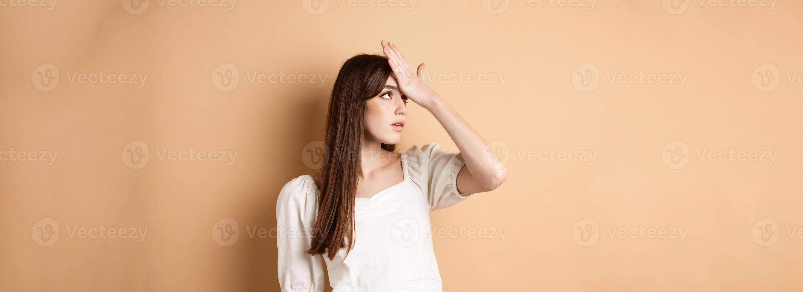 Girl rolling her eyes and slap forehead annoyed, bothered by something stupid or lame, making face palm on beige background photo