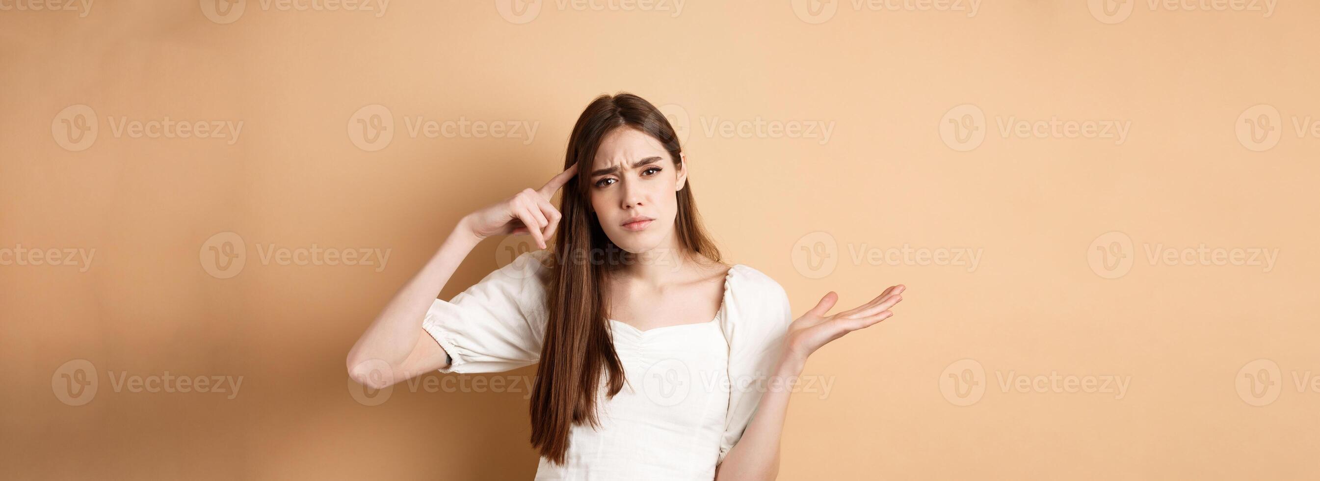 Are you stupid. Annoyed woman mocking person, pointing finger at head and shrugging confused, irritated by crazy ideas, standing on beige background photo