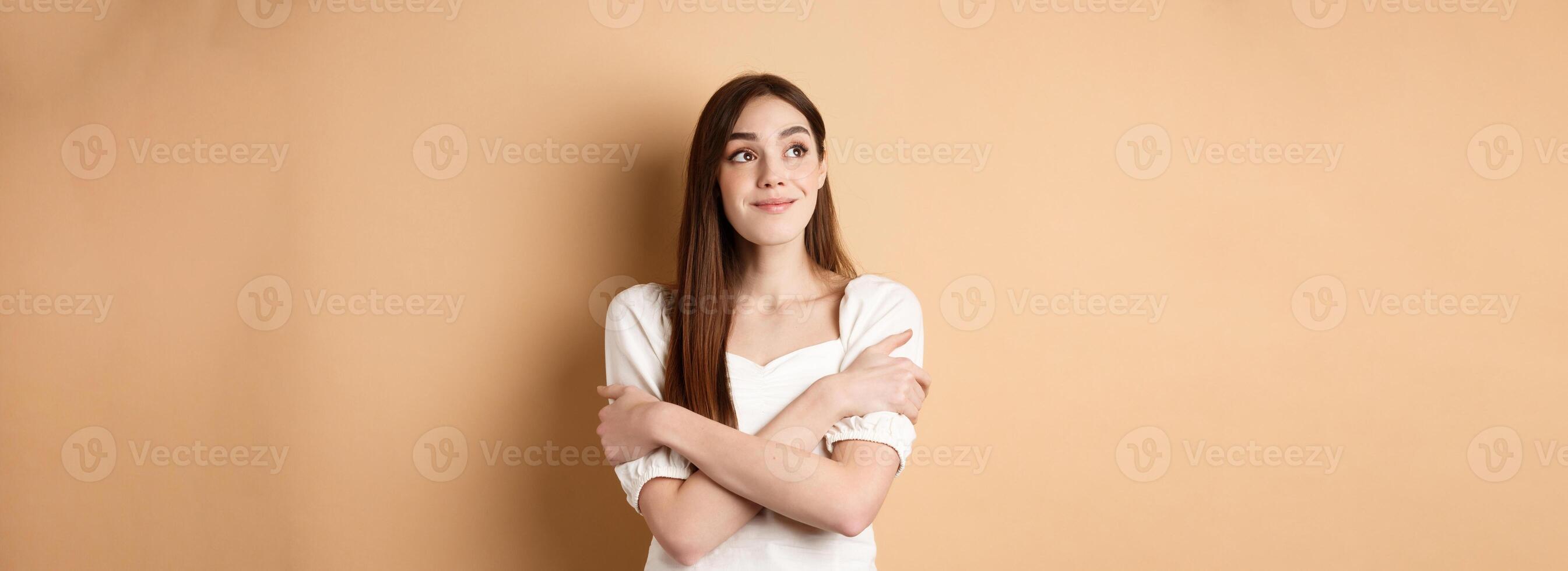 Romantic young girl dreaming, hugging herself and smiling at upper left corner, imaging something lovely, standing on beige background photo
