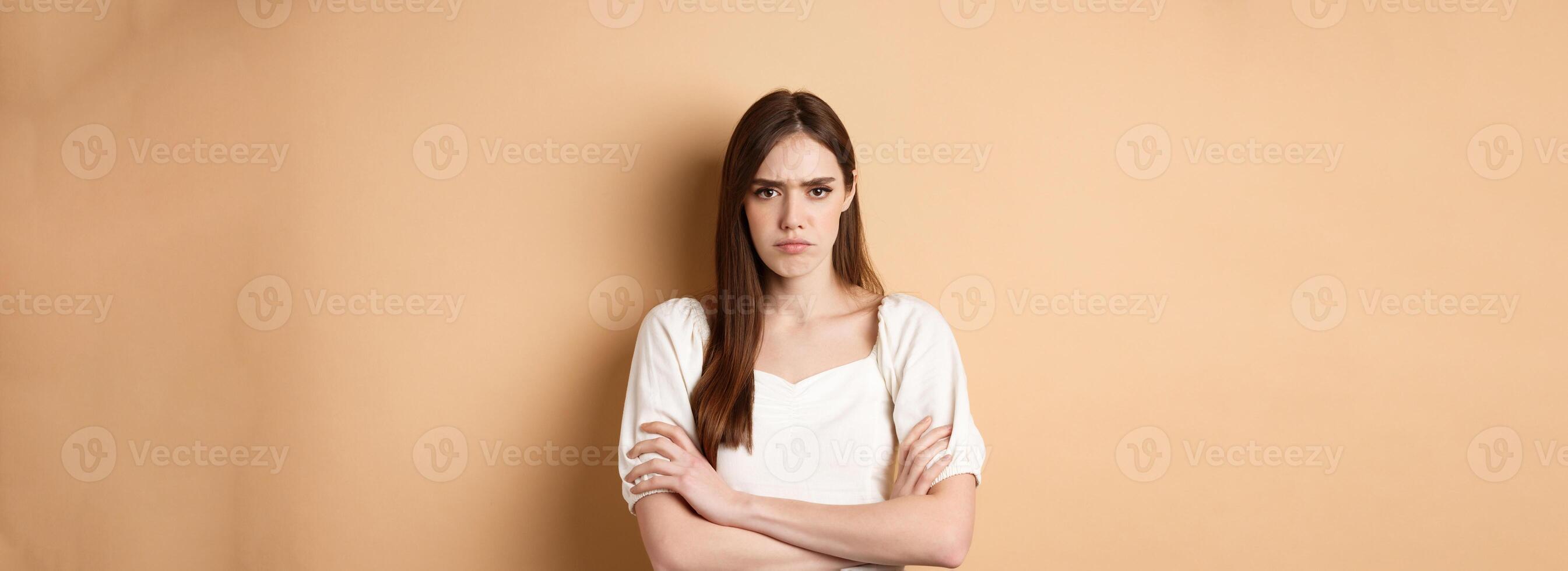 Angry girlfriend cross arms on chest and frowning, looking offended or disappointed, sulking at you, standing mad on beige background photo