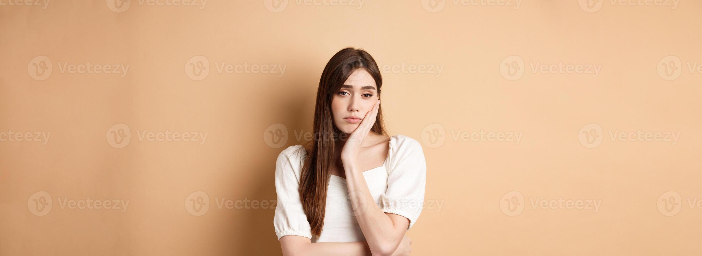 Bored and tired young woman looking indifferent at camera, lean face on palm and stare at camera unamused, standing sleepy on beige background photo