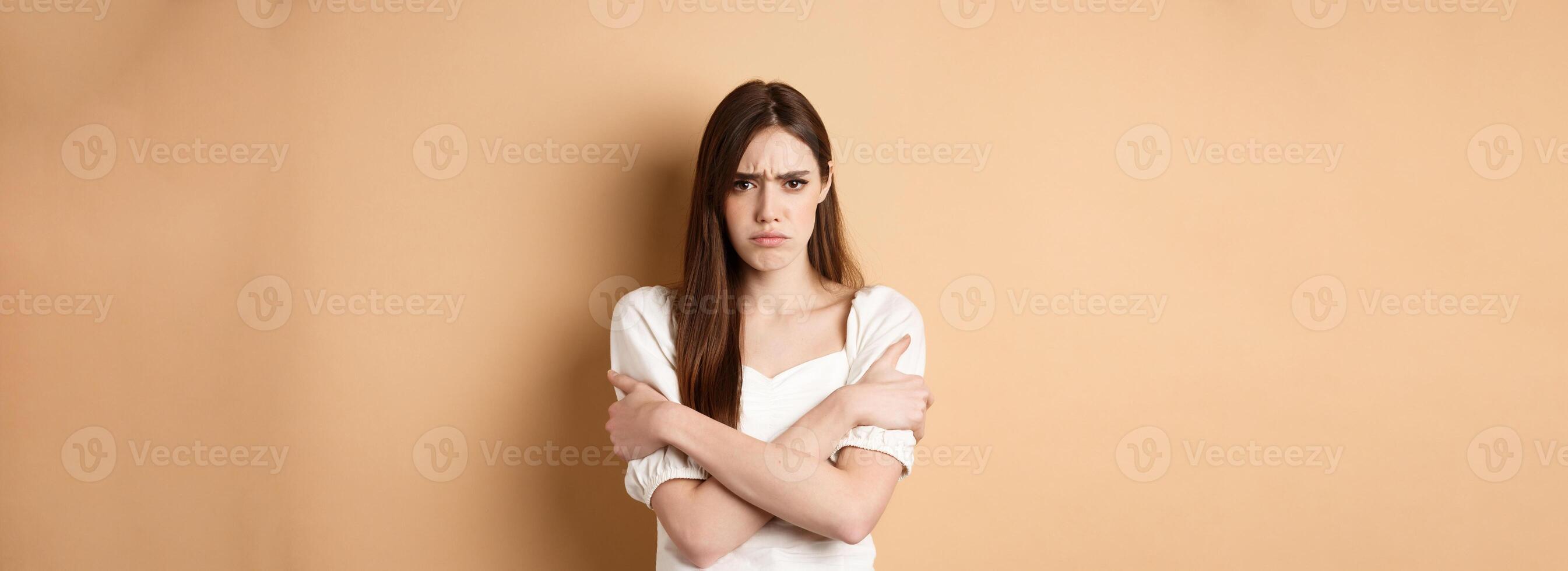 Offended woman hugging herself and frowning upset, feeling timid and defensive, standing on beige background photo