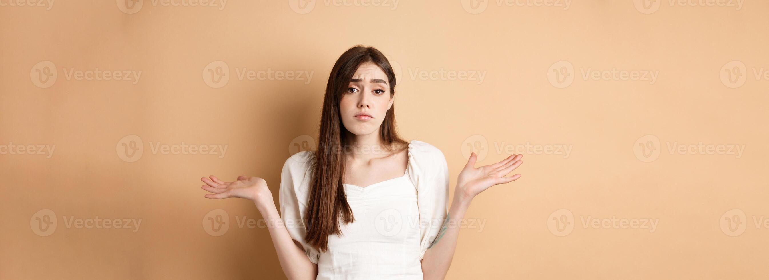 I dont know. Clueless sad girl shrugging shoulders and looking unaware, have nothing to say, standing on beige background photo
