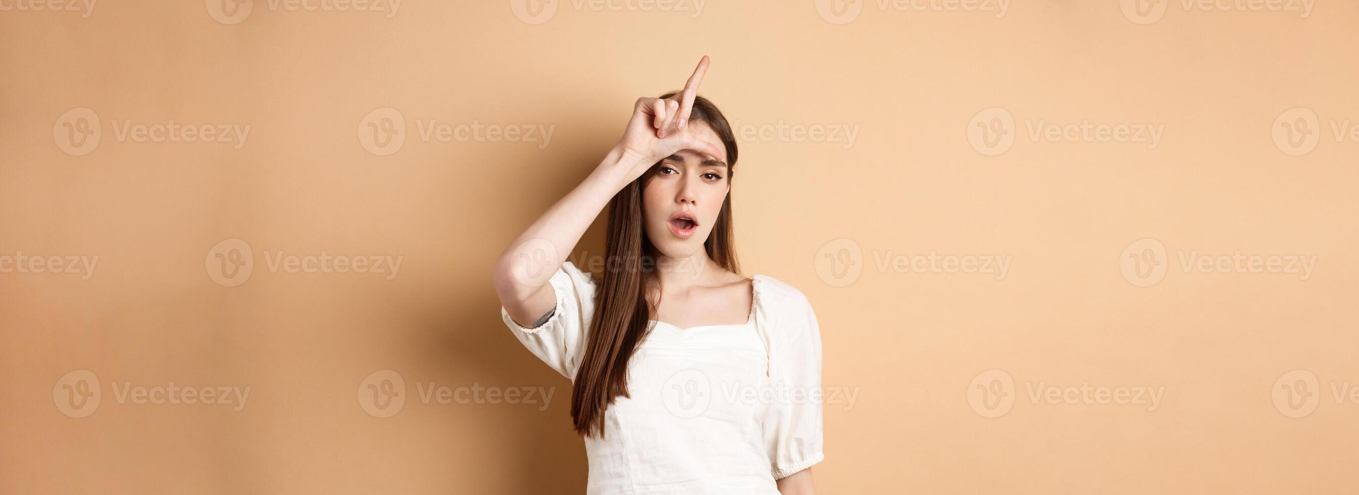 Confident woman mocking people with loser sign, being mean, standing on beige background photo