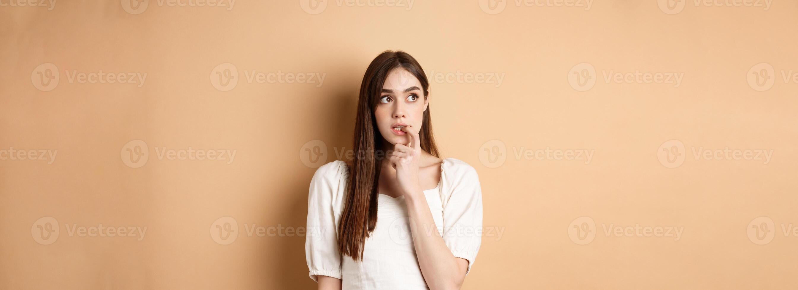 Nervous young girl biting fingernail and looking aside excited, making choice, thinking about something, standing on beige background photo