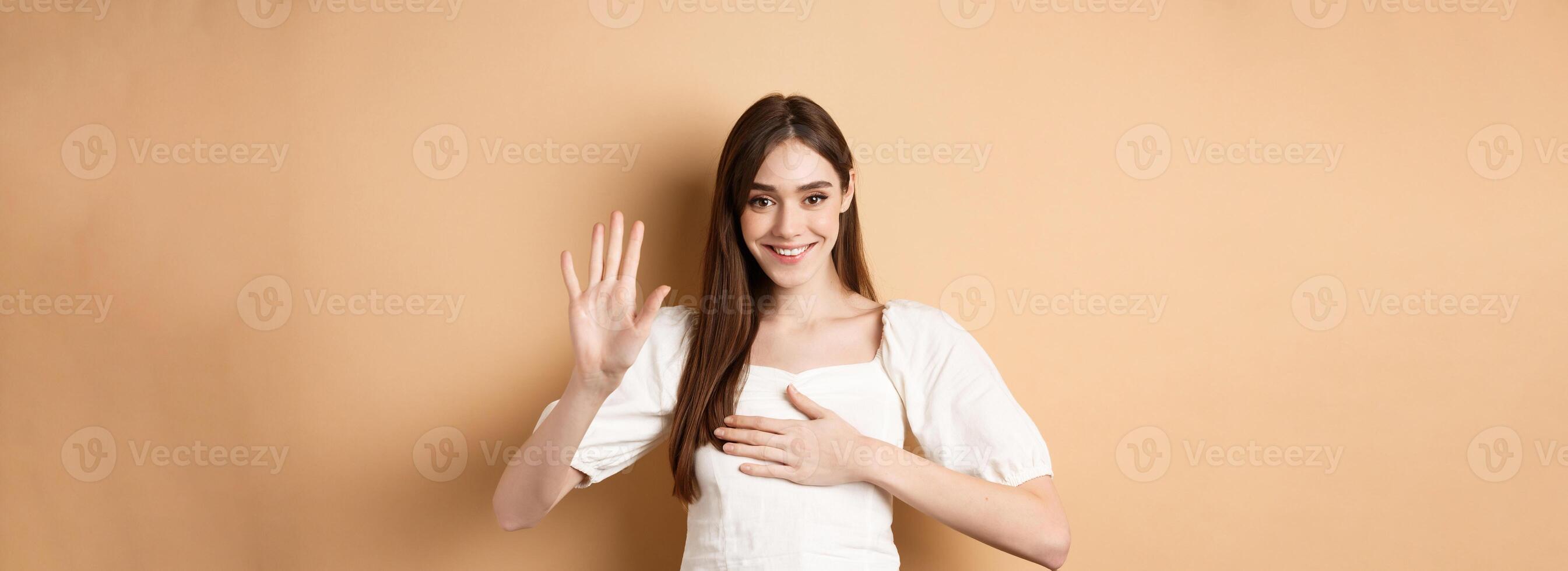 Cute smiling girl make promise, put hand on heart and tell truth, being honest, swearing to you, standing on beige background photo