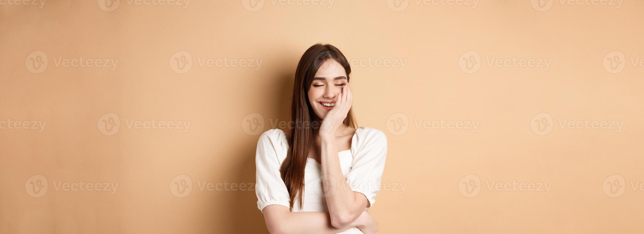 Happy and romantic girl laughing with eyes closed, touching face and feeling joy, standing on beige background photo