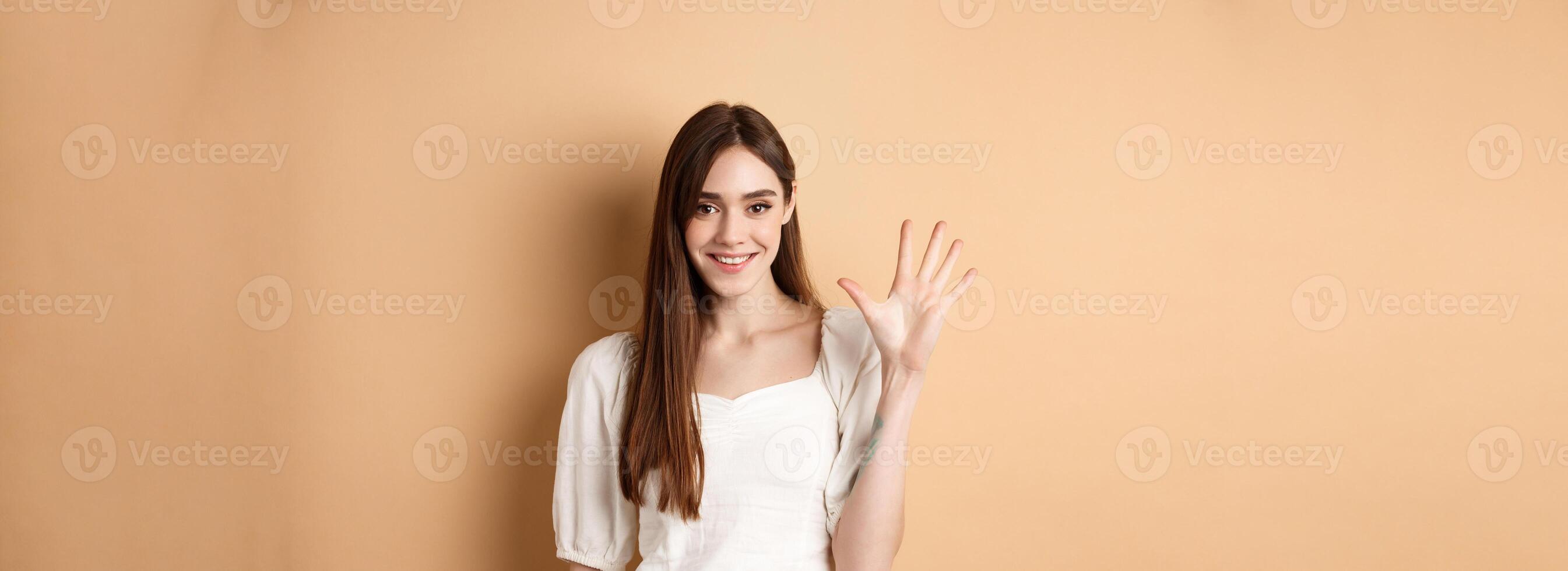 Attractive young woman show fingers number five, smiling and looking confident, standing on beige background photo