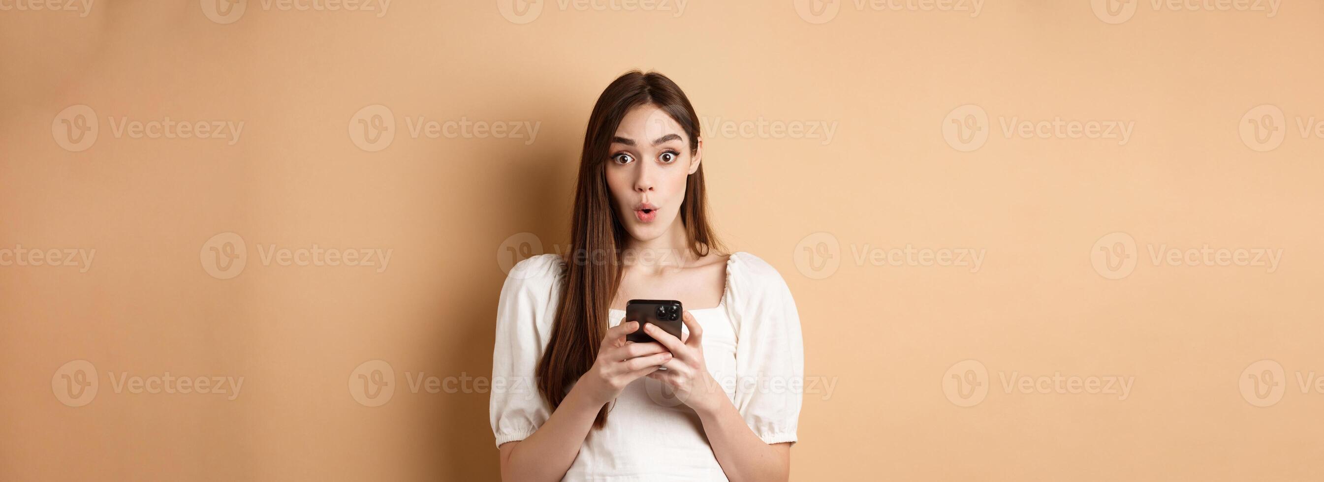 Girl standing amazed with smartphone, saying wow and look excited by awesome online offer news, standing on beige background photo