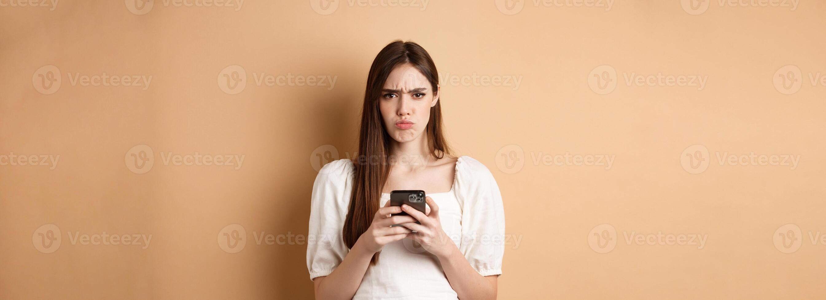 Disappointed girl with smartphone frowning, pucker lips upset, reading bad news on phone, standing on beige background photo