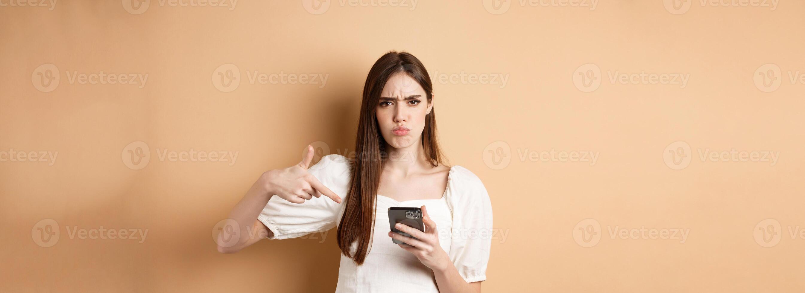 Online shopping concept. Angry frowning girl pointing at mobile phone screen, look disappointed, standing on beige background photo
