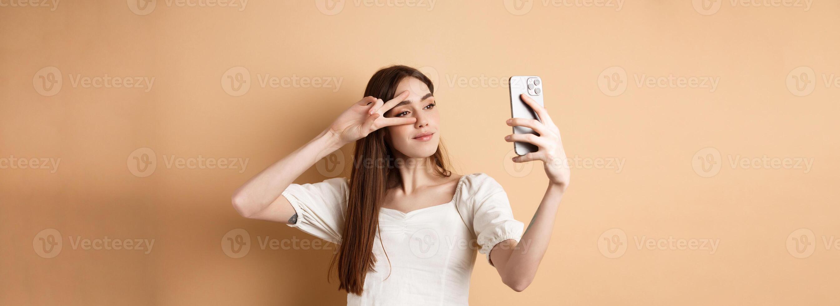 Cute woman taking selfie on smartphone, showing v-sign near eye, posing for social media photo, standing on beige background photo