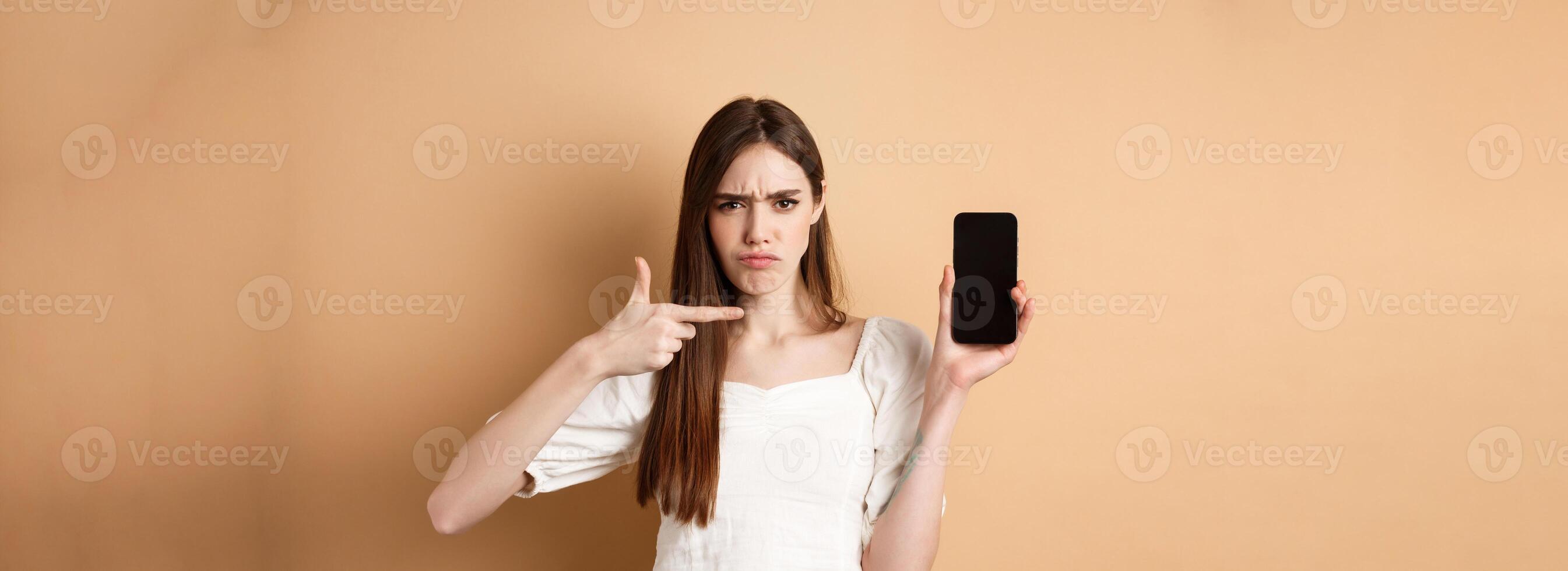 Disappointed frowning girl pointing at empty phone screen, complaining at online news, standing on beige background photo