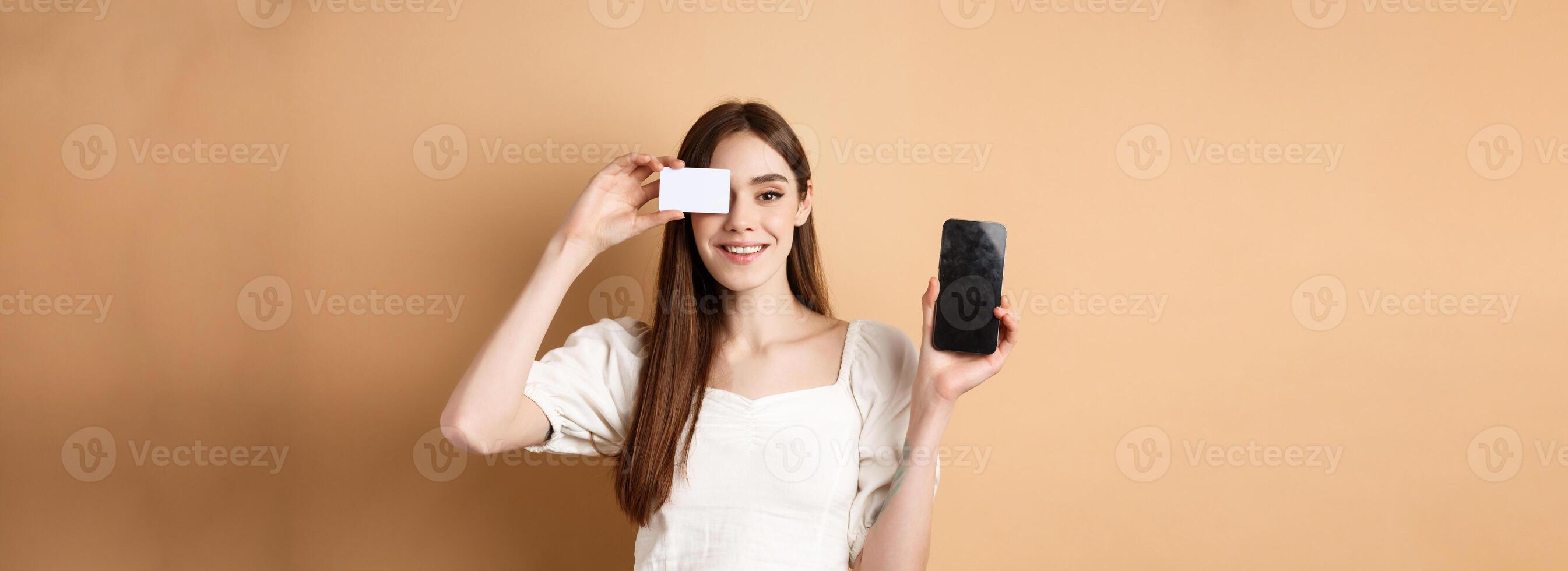 Cheerful young woman showing plastic credit card and empty mobile phone screen, smiling pleased at camera, standing on beige background photo