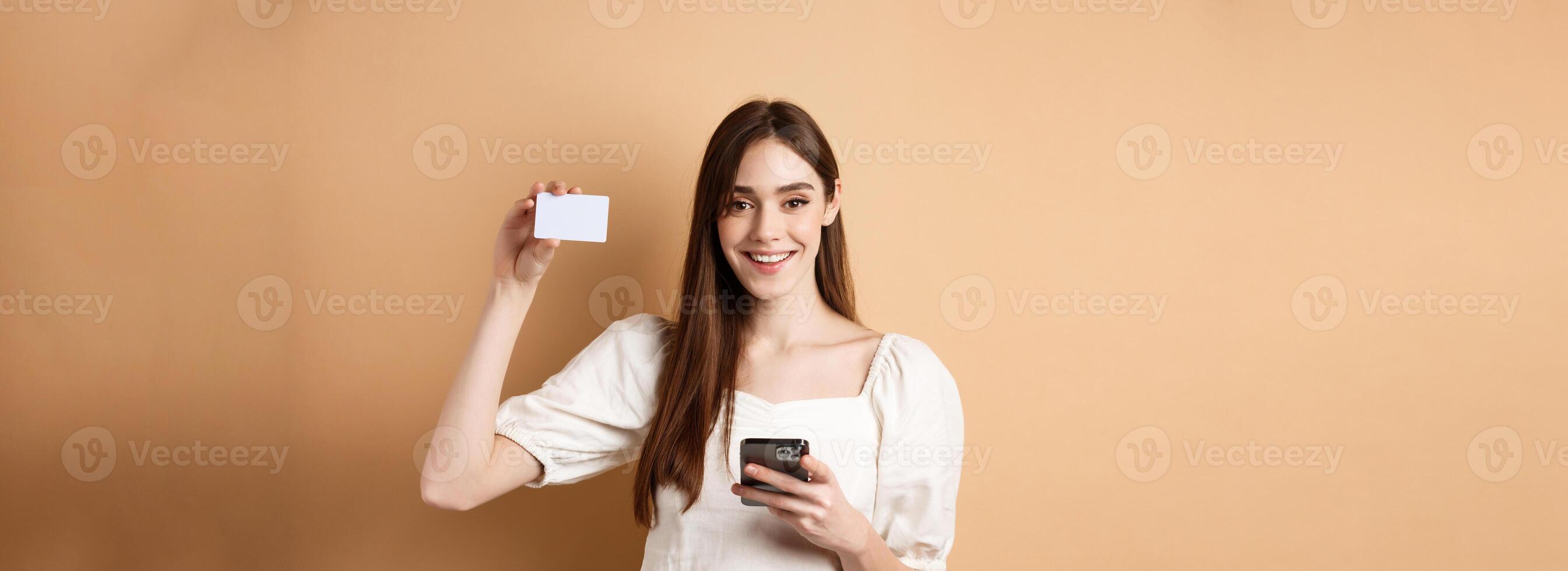 E-commerce concept. Smiling woman showing plastic credit card while shopping online on mobile phone, standing against beige background photo