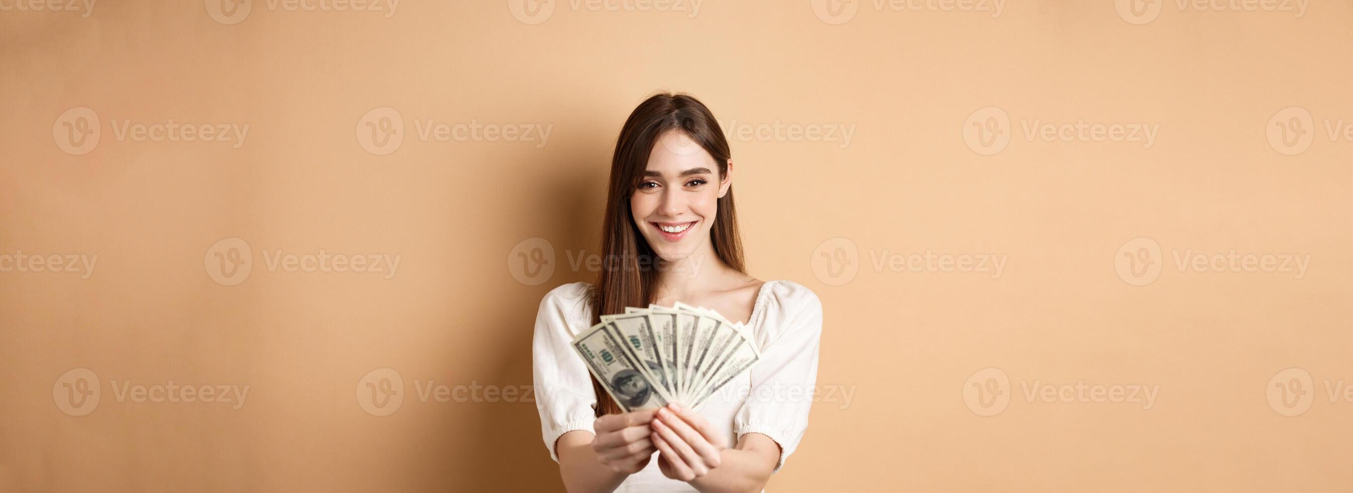 Young woman showing dollar bills and smiling, standing with money on beige background. Concept of loan and insurance photo