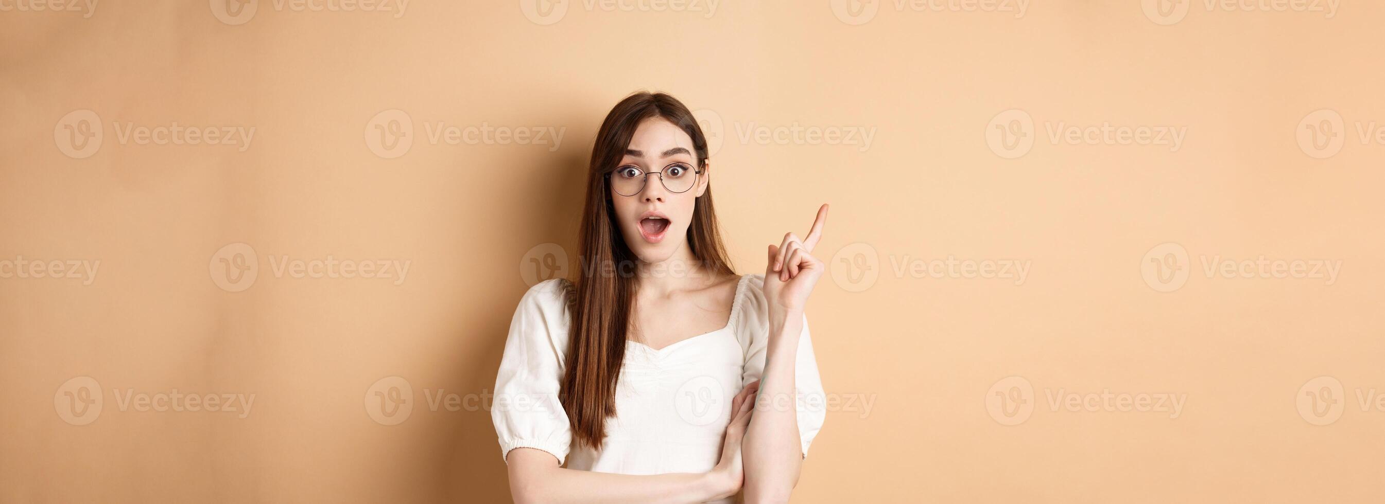 Creative young woman in glasses pitching idea, raising finger in eureka sign and gasping, have a plan, standing on beige background photo
