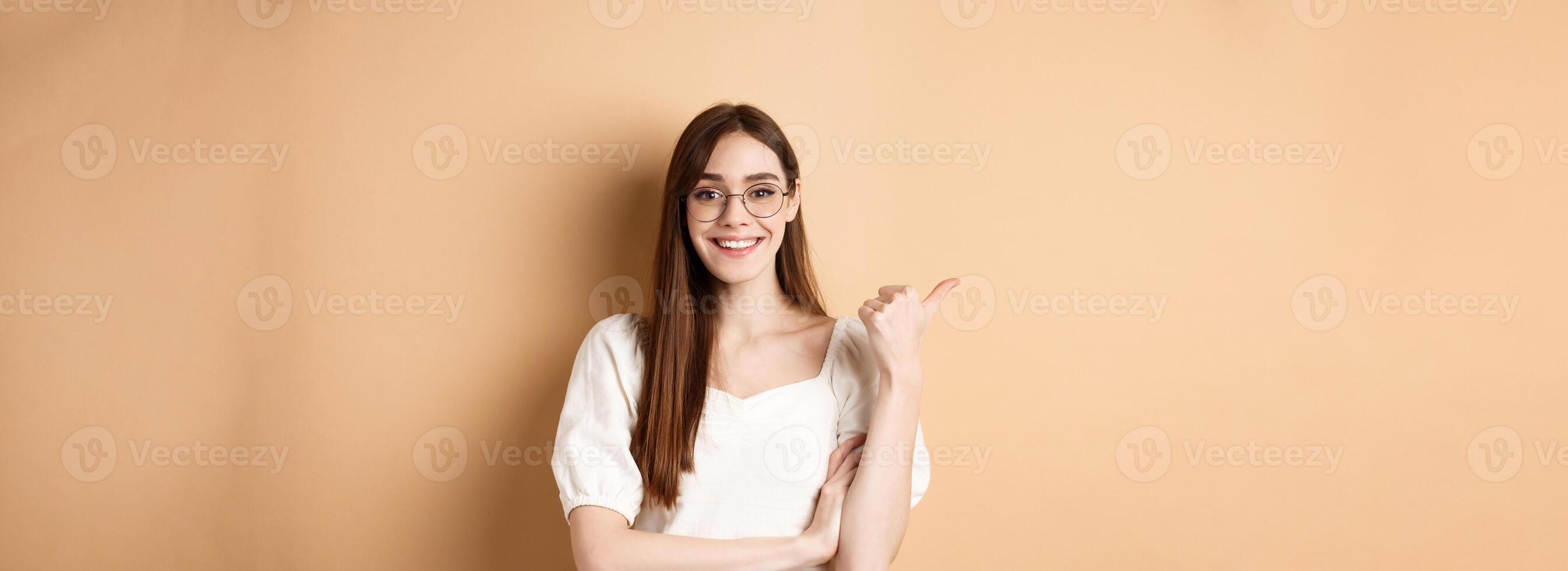 Cheerful woman in eyewear pointing finger left at logo, smiling happy, standing on beige background photo