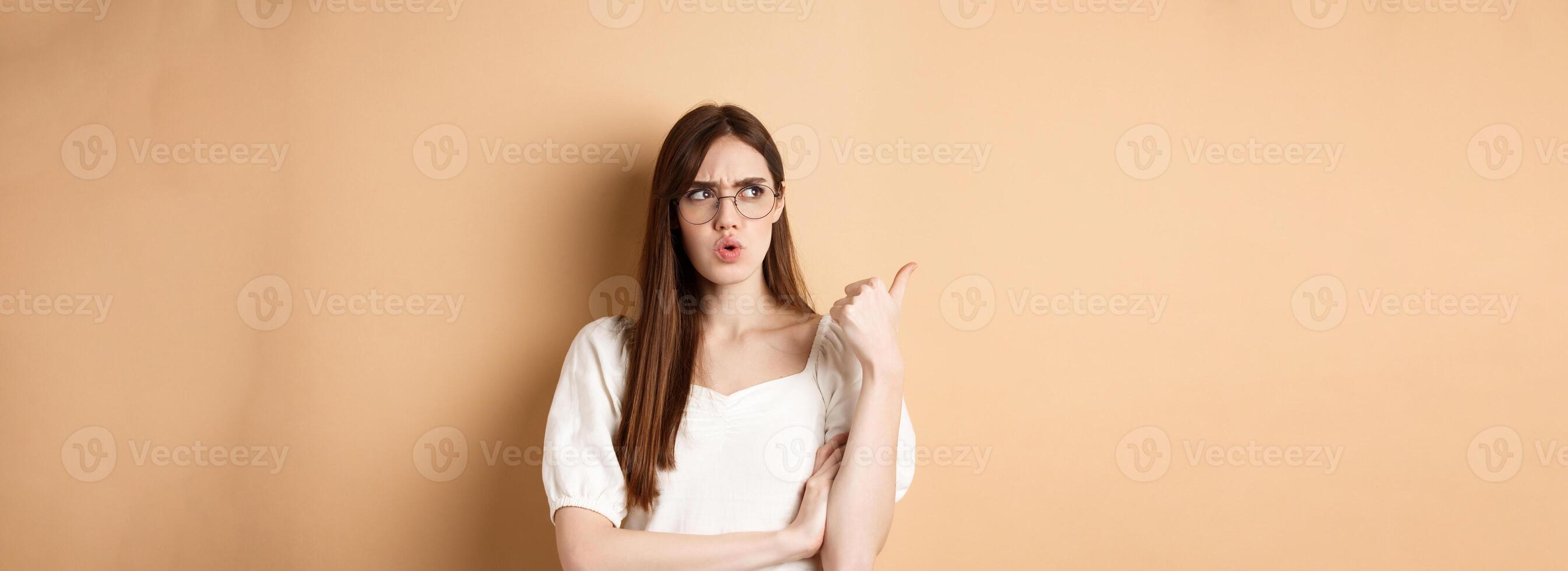 Confused and suspicious young woman in glasses frowning, pointing and looking aside with displeased face expression, standing on beige background photo