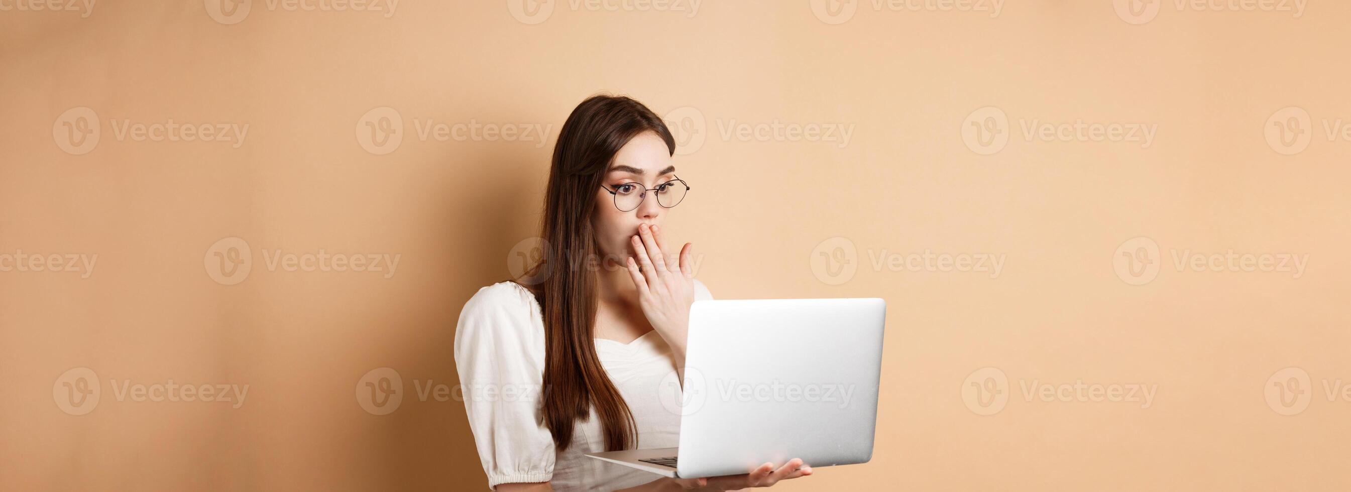 Shocked girl look at laptop screen, covering mouth with amazed face, standing in glasses on beige background photo