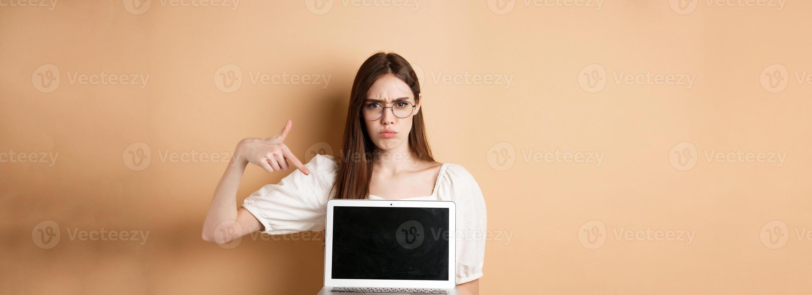Angry frowning woman in glasses pointing at laptop screen, complaining and look disappointed, standing on beige background photo