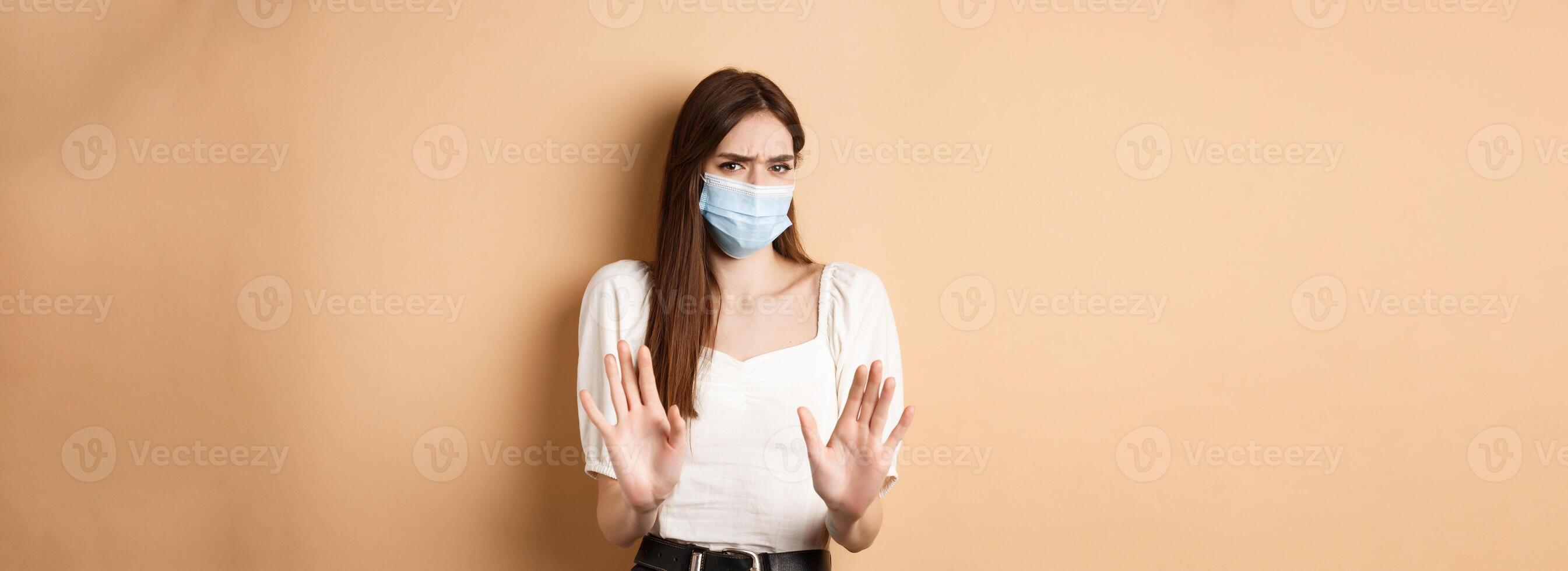 Pandemic and healthcare concept. Keep distance. Young woman in medical mask stretch out hand in stop sign, telling to stay away and frowning disgusted, beige background photo