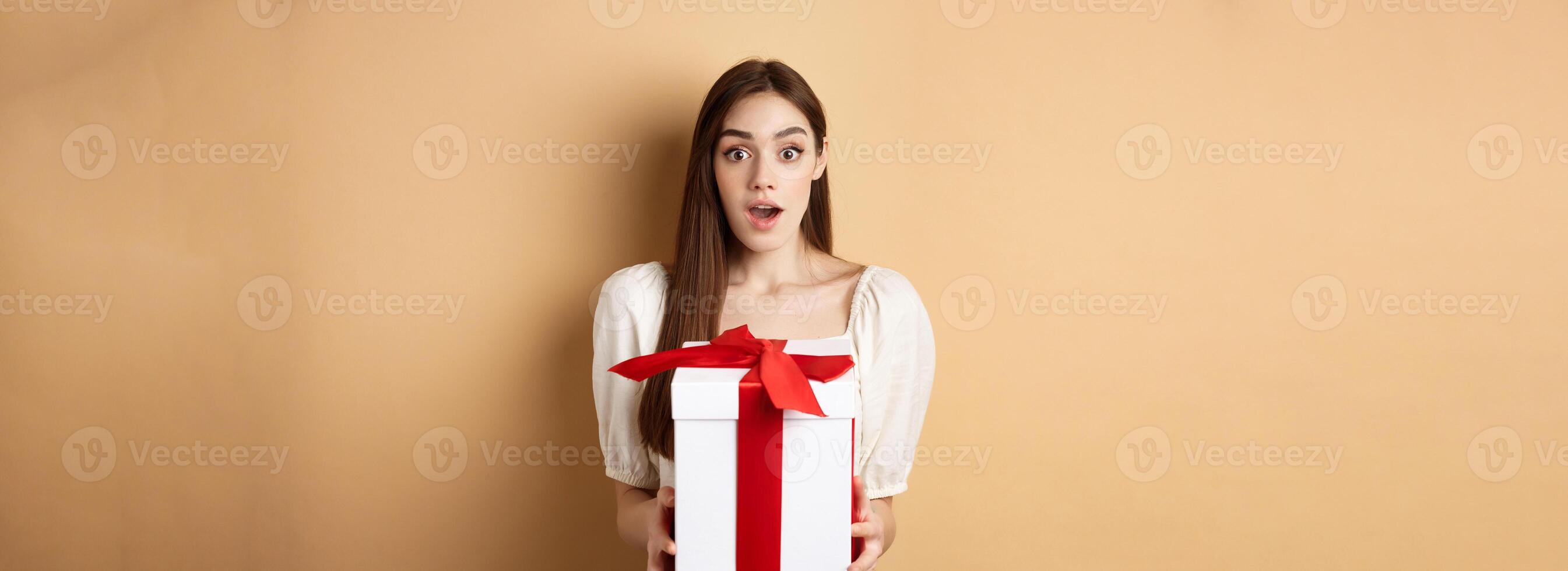 Amazed beautiful girl receiving gift from secret admirer on Valentines day, looking surprised at camera, holding box and standing on beige background photo