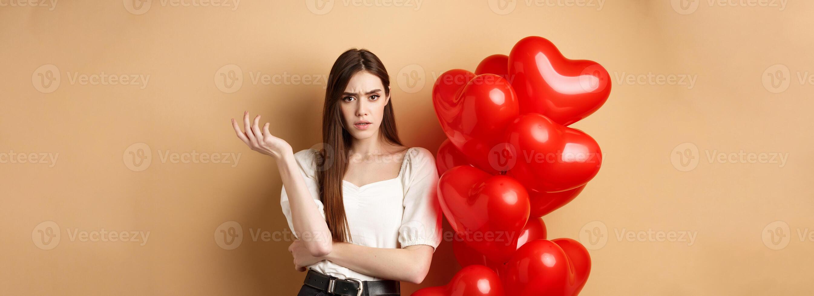 Valentines day and love concept. Confused girlfriend having argument with lover, raising hand and looking annoyed at camera, cant understand something, standing near hearts balloons photo