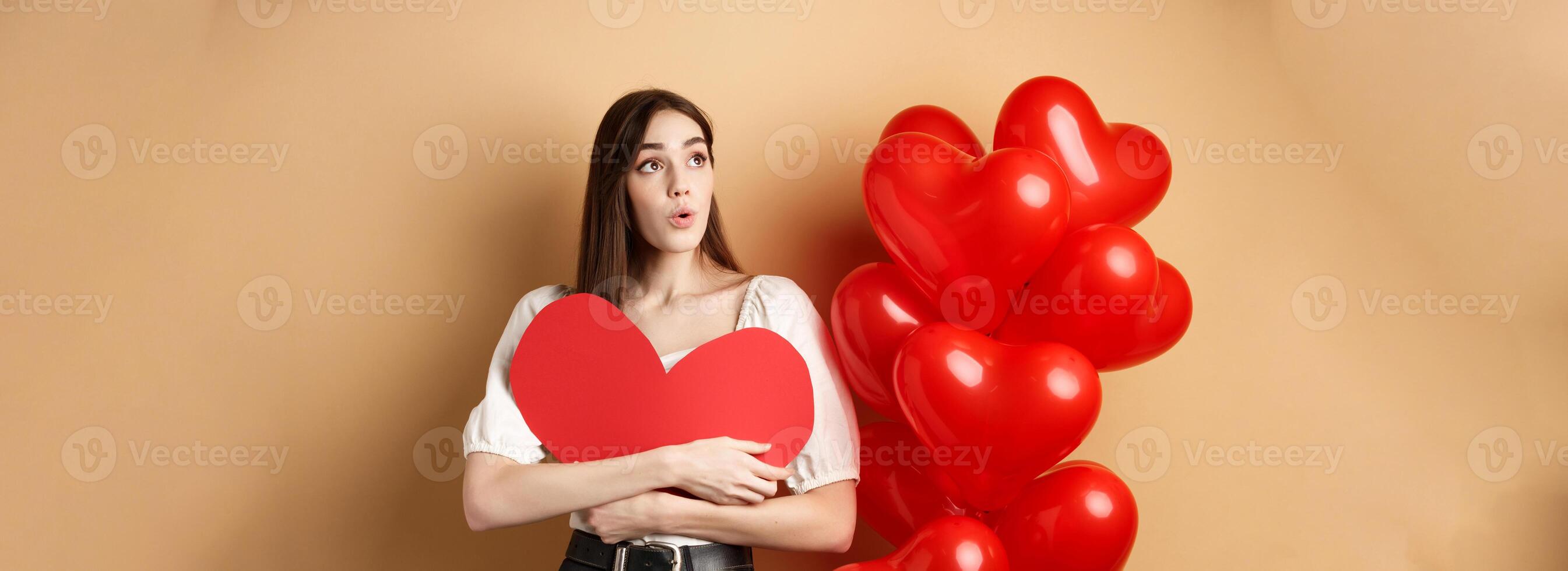 Valentines day and love concept. Intrigued tender girl hugging big red heart cutout and stand near balloons, looking aside at logo, saying wow, beige background photo