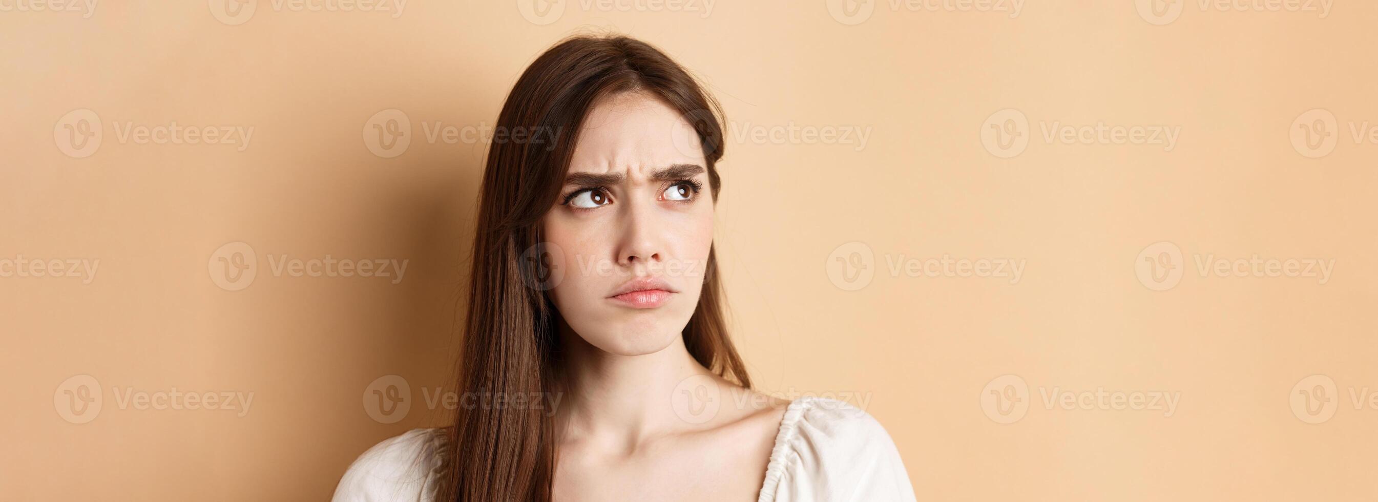 Close up of angry cute girl frowning, looking aside displeased, stare at bad thing, standing on beige background photo