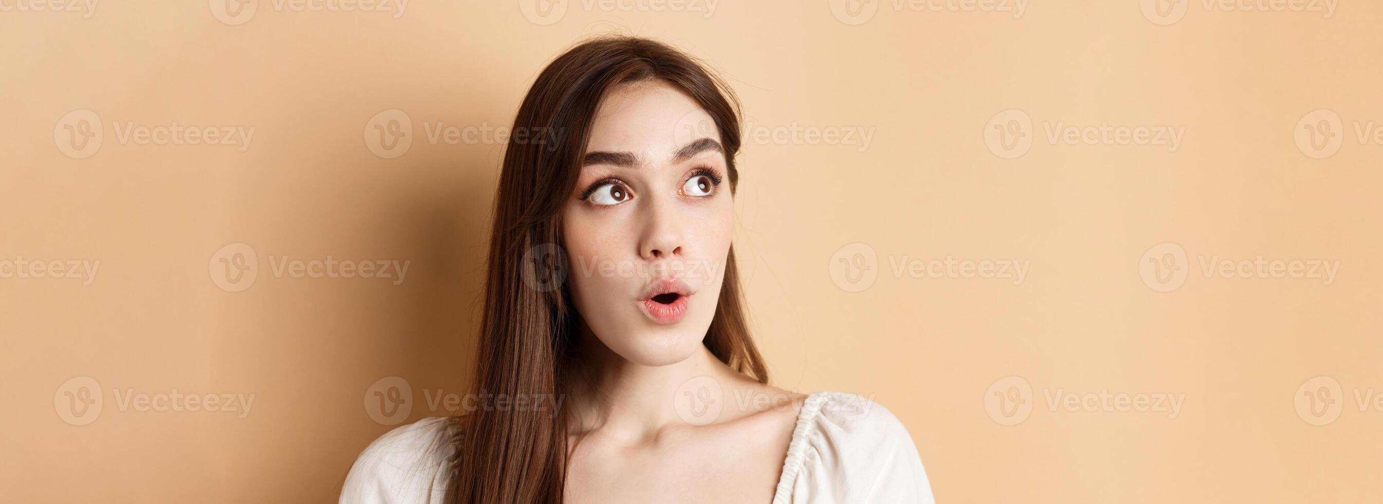 Close up portrait of excited pretty woman looking aside logo, say wow with amazed face, standing on beige background photo