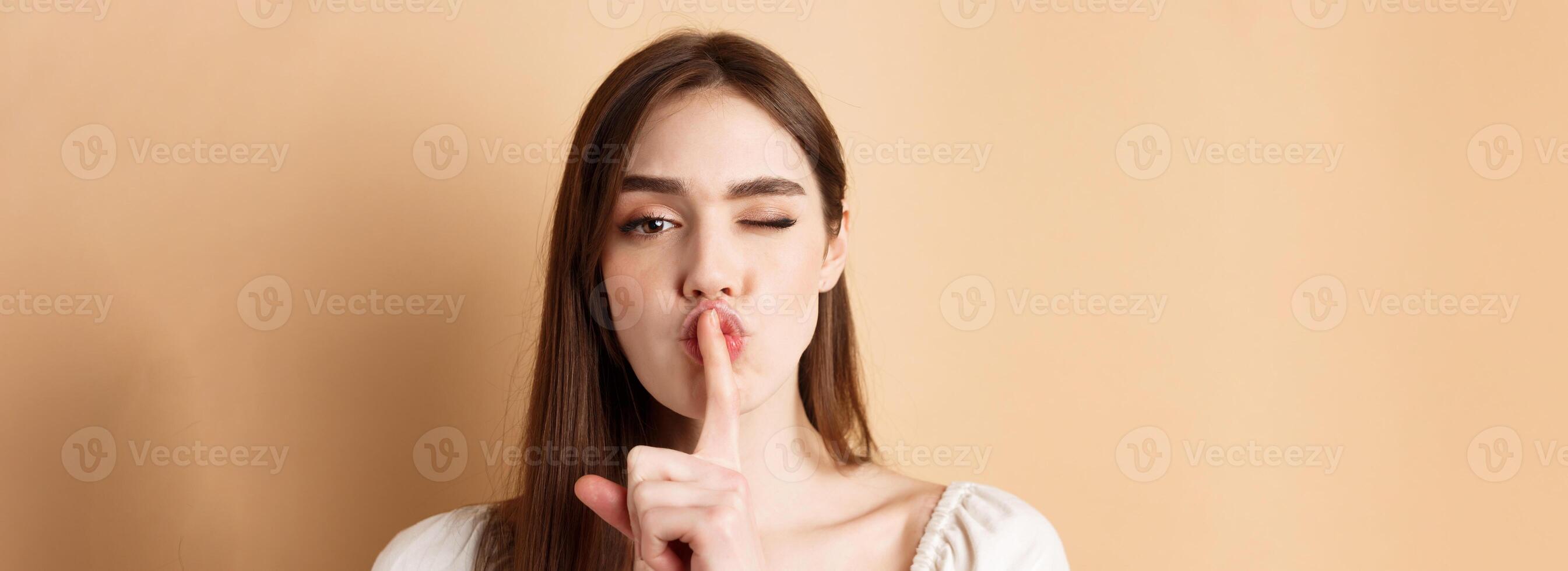 Close up of attractive woman shushing at camera, hiding secret, making hush sign and winking, show taboo sign, standing on beige background photo
