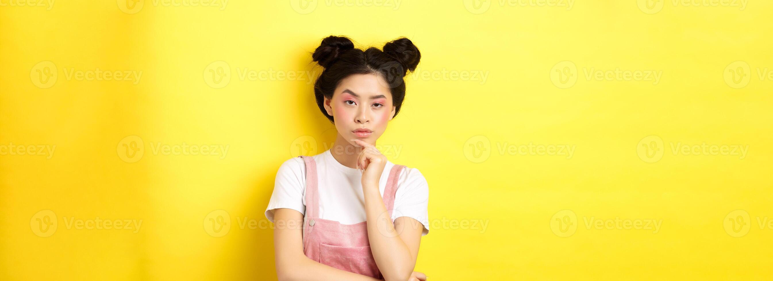 Suspicious asian girl looking at camera and thinking, stare with disbelief, standing with makeup on yellow background photo