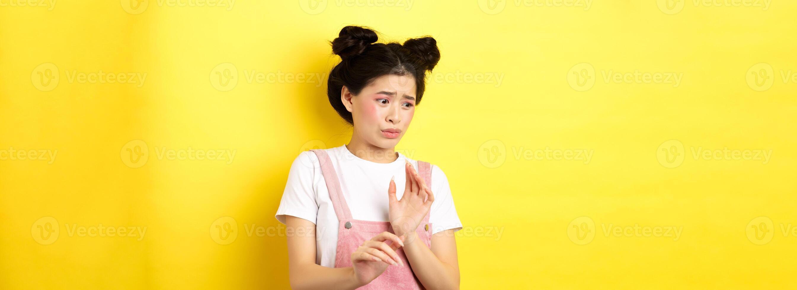 Disgusted asian girl raising hands up defensive, block something disgusting, turn away from aversion and reluctance, standing on yellow background photo