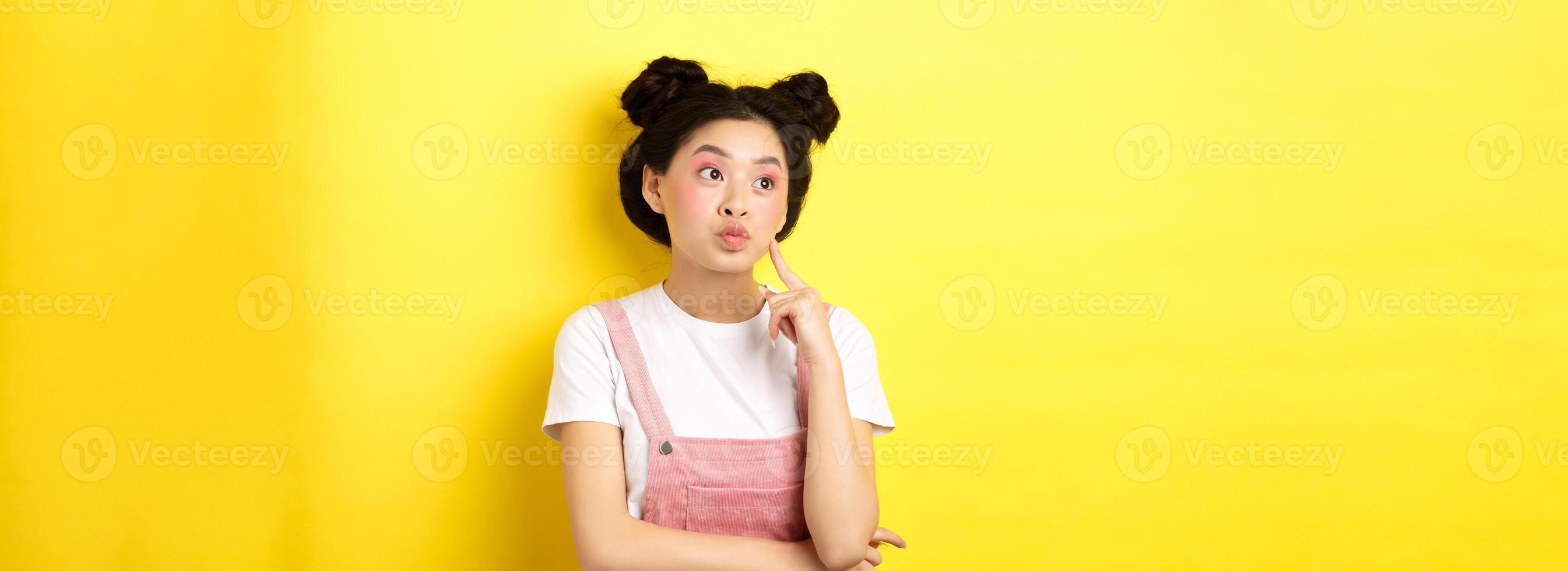 Summer lifestyle concept. Pensive glamour asian girl thinking, looking left at logo and dreaming, making choice, standing on yellow background photo