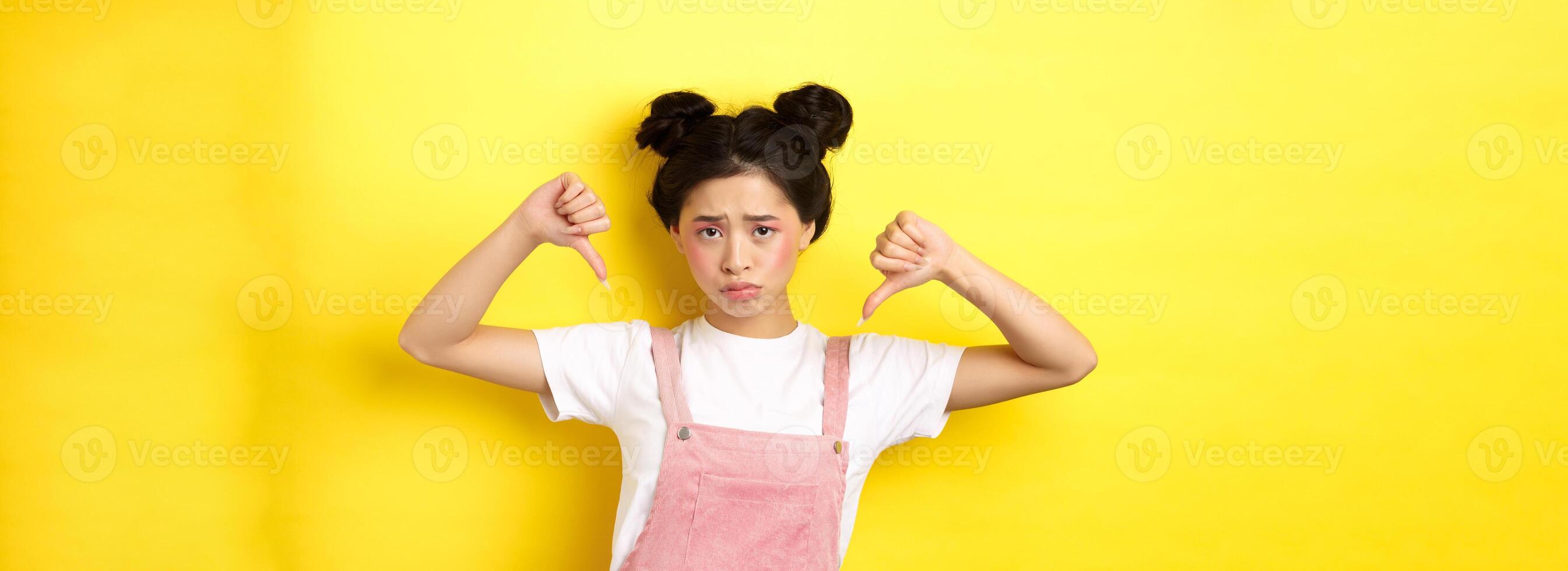 Summer lifestyle concept. Sad and gloomy asian woman with makeup, pointing fingers down and frowning upset, disappointed with unfair situation, yellow background photo