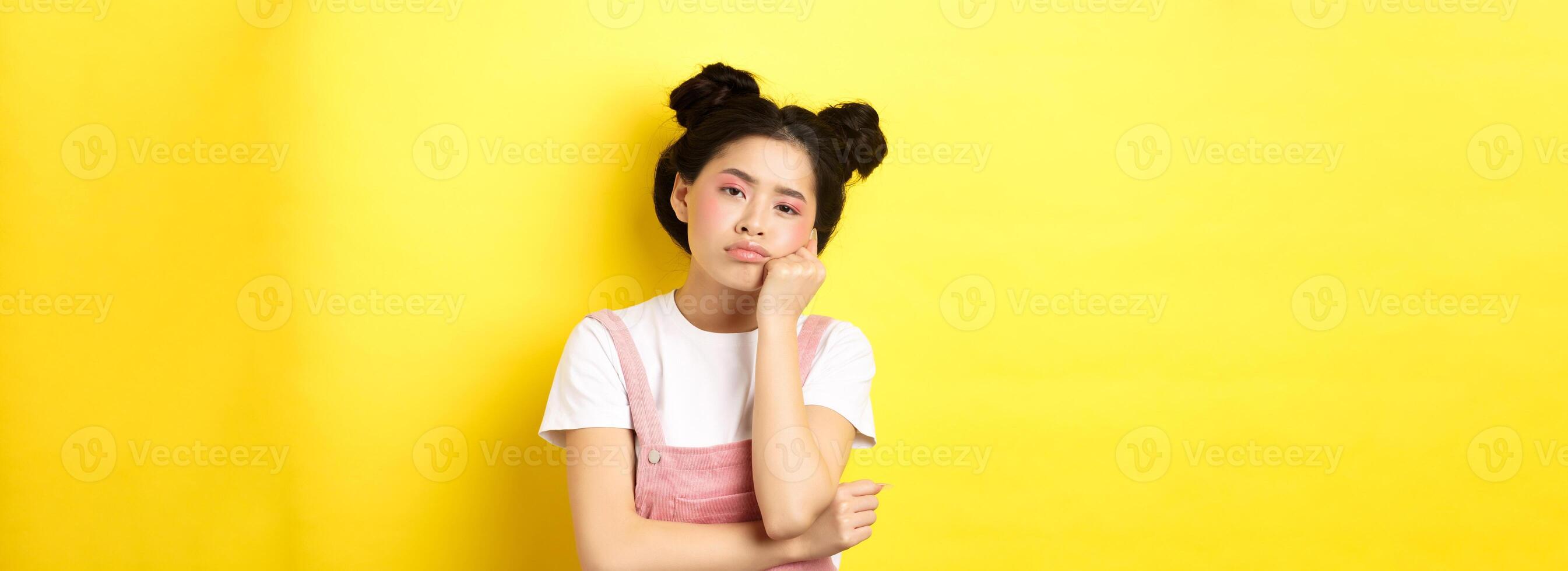Sad and bored teen asian girl standing alone, looking at camera indifferent, staring with boredom, yellow background photo