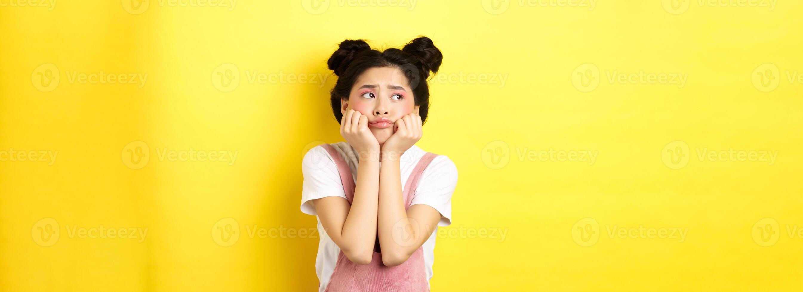 Sad moody asian girl, lean face on hands, looking left with sulking expression, standing in summer clothes on yellow background photo