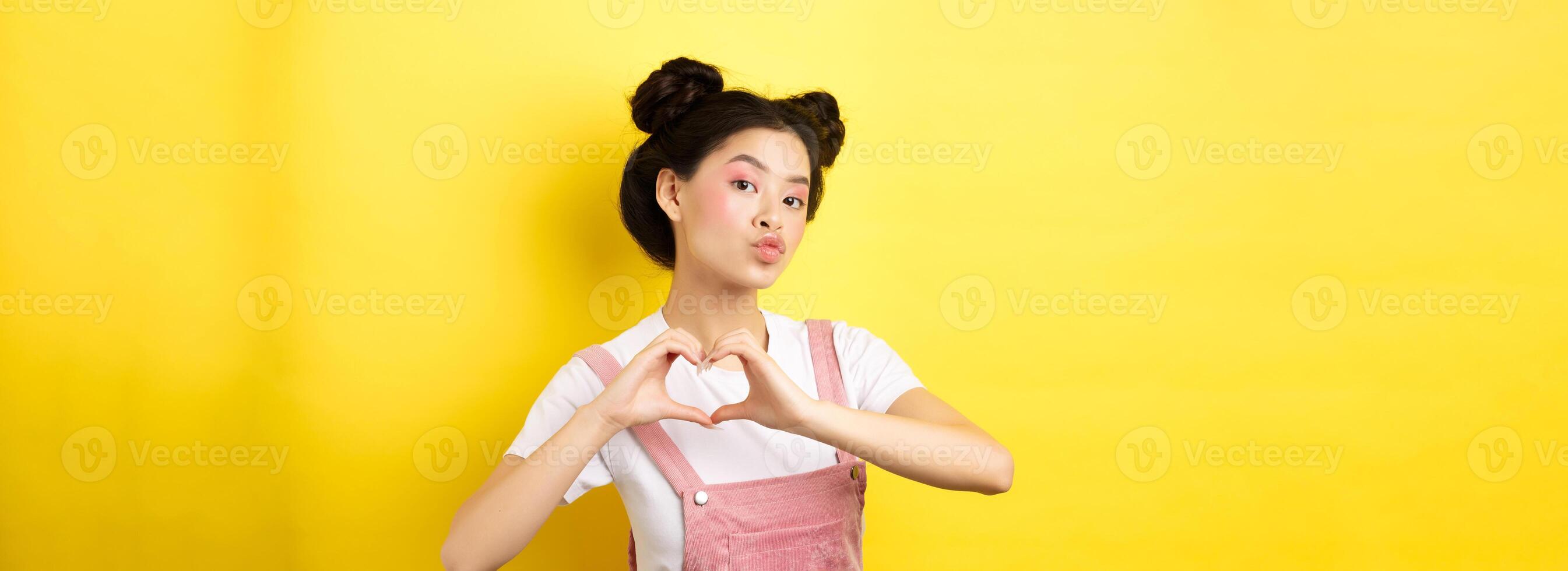 Silly japanese girl showing heart gesture and wishing happy Valentines day, pucker lips to kiss lover, standing on yellow background photo