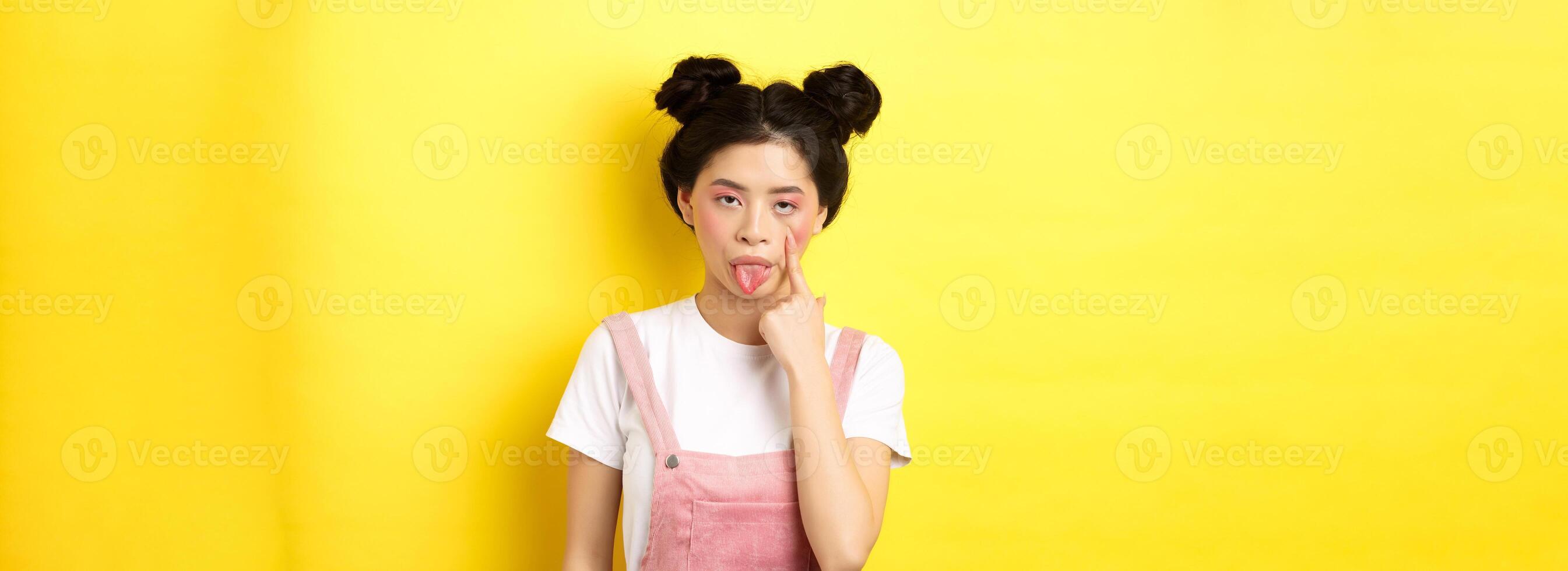 Rude asian girl stretch eyelid and showing tongue, mocking someone, standing on yellow background photo