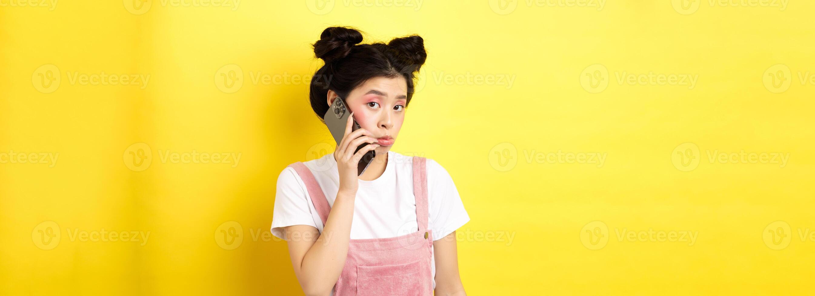 Cute teen girl talking on smartphone, making silly pouting face and look timid at camera, standing with glamour makeup on yellow background photo