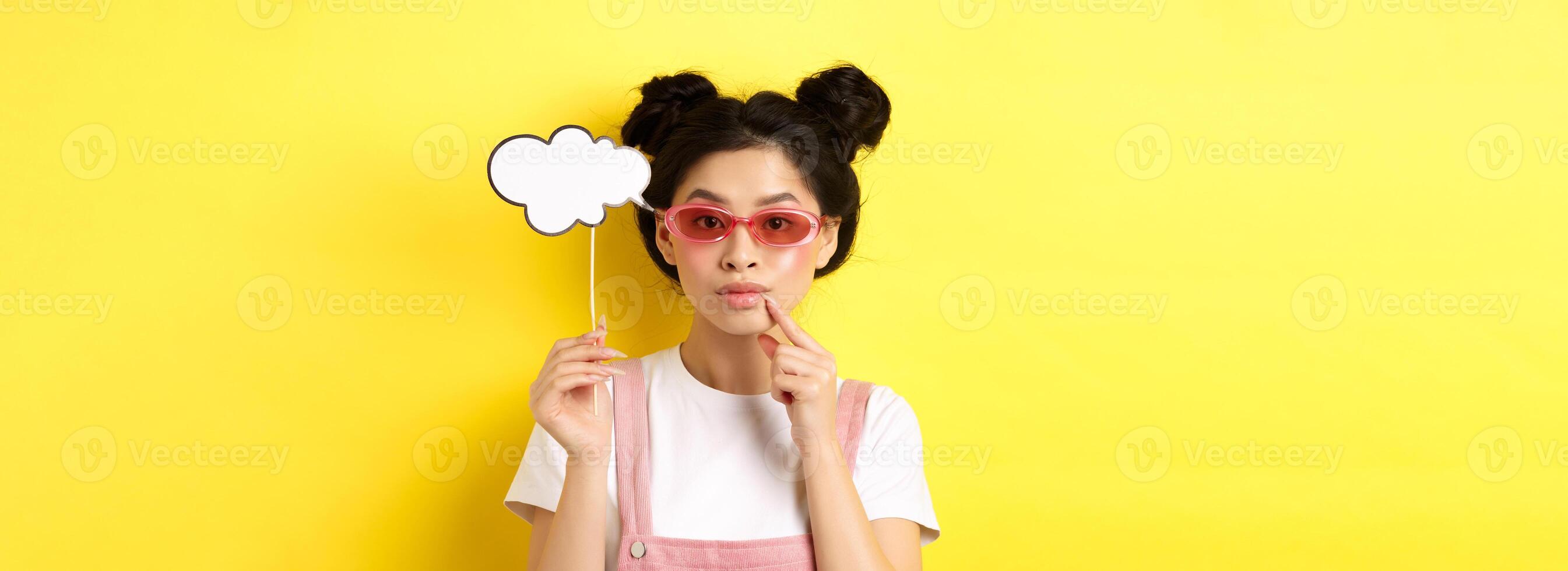 Summer and fashion concept. Stylish asian girl in sunglasses holding comment cloud on stick and looking thoughtful, thinking or saying something, standing on yellow background photo