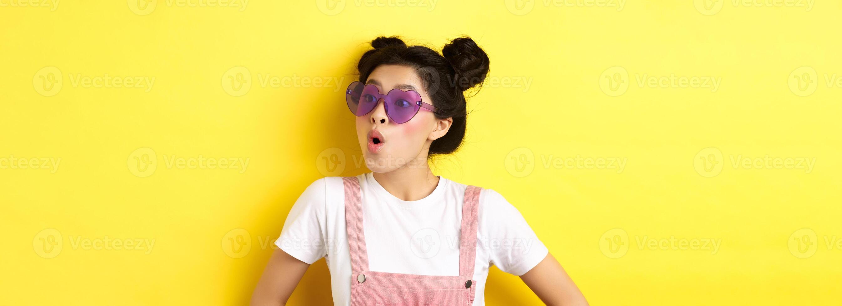 Summer and fashion concept. Close-up of surprised asian teen girl in heart-shaped sunglasses looking right side with excited face, standing on yellow background photo