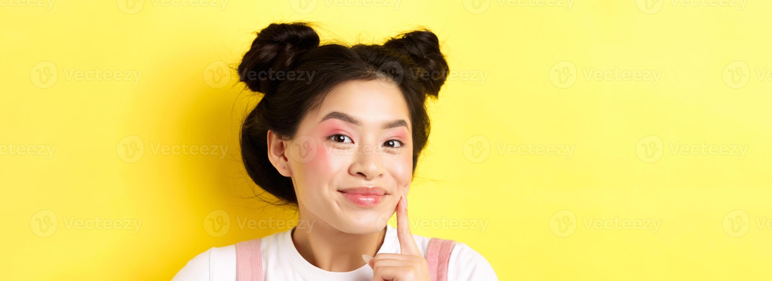 Beauty. Stylish asian girl with bright glamour makeup, touching soft and shiny facial skin, smiling happy at camera, standing on yellow background photo