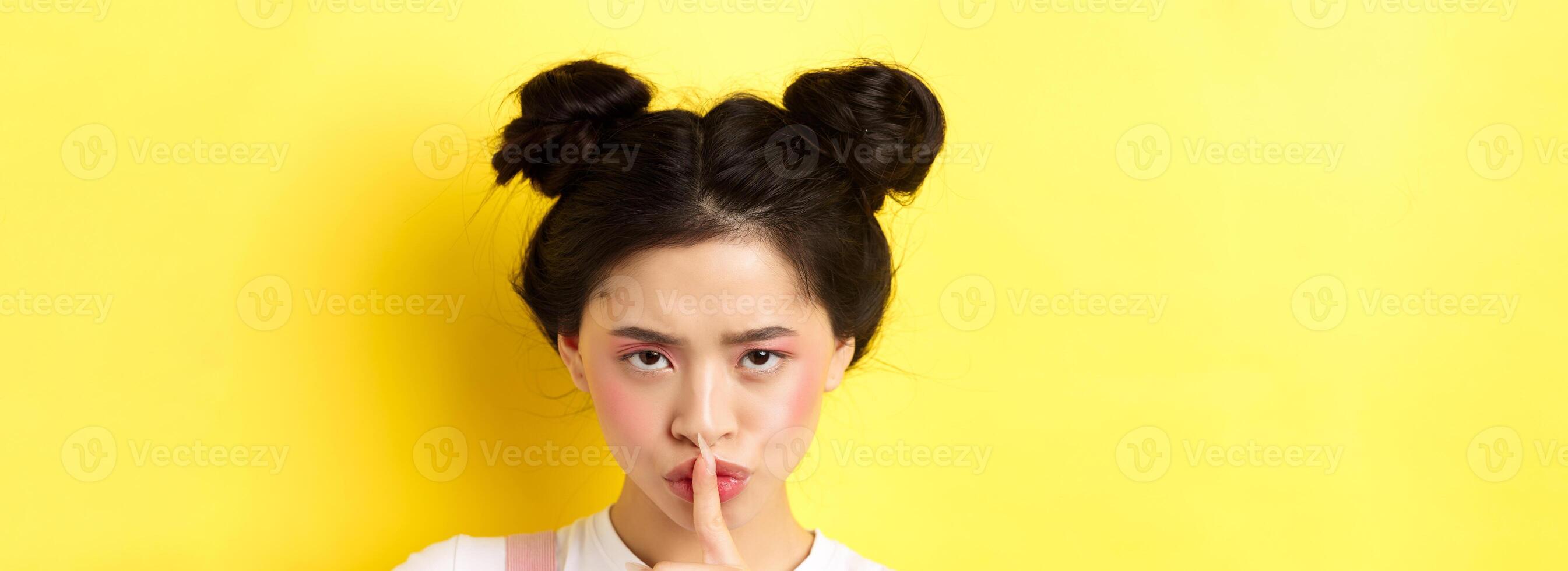 Close up portrait of angry asian girl shushing, press fingers on lips and tell be quiet, scolding loud person, need silence, telling big secret, standing on yellow background photo